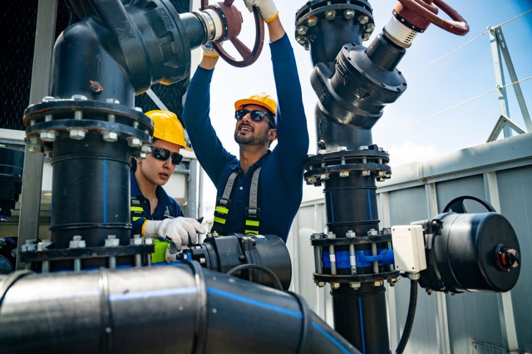 Two workers in hard hats inspect industrial pipes and valves at a power plant.