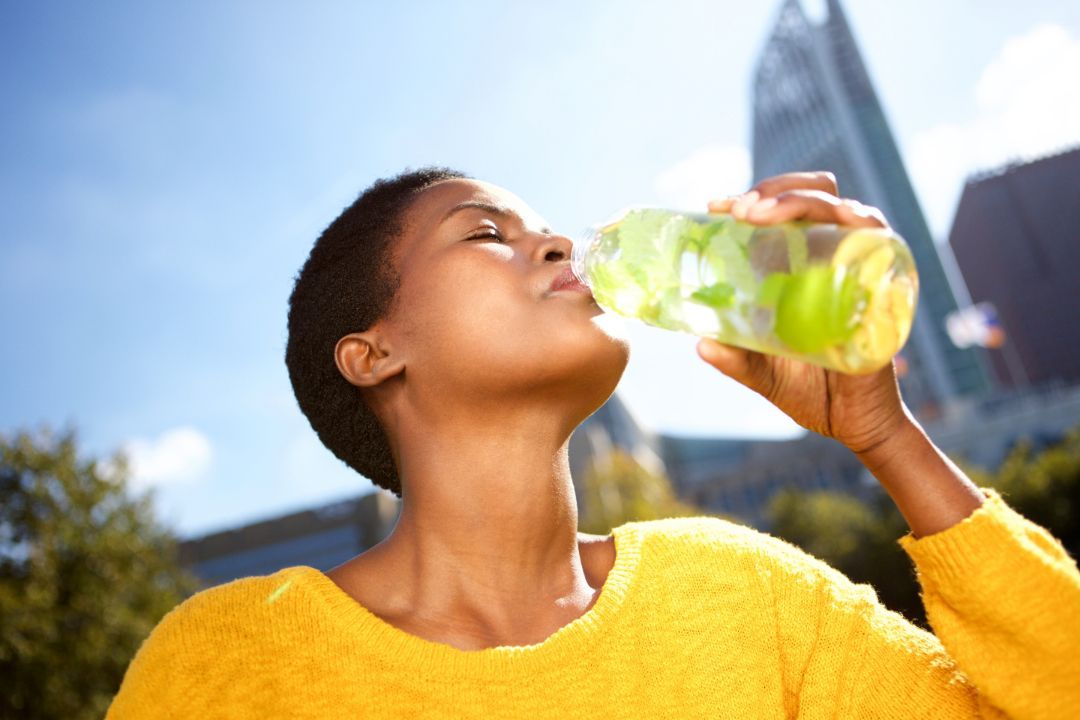 Person in a yellow sweater drinking from a green water bottle outdoors in bright sunlight