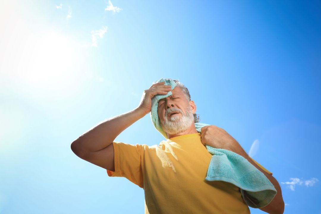 Man wiping sweat with a towel under a bright blue sky, looking overheated