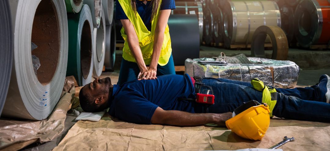 A person in a high-visibility vest performs CPR on an unconscious worker lying on a warehouse floor near metal coils.