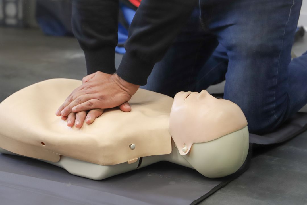 A person kneels on a mat, performing chest compressions on a CPR training mannequin.