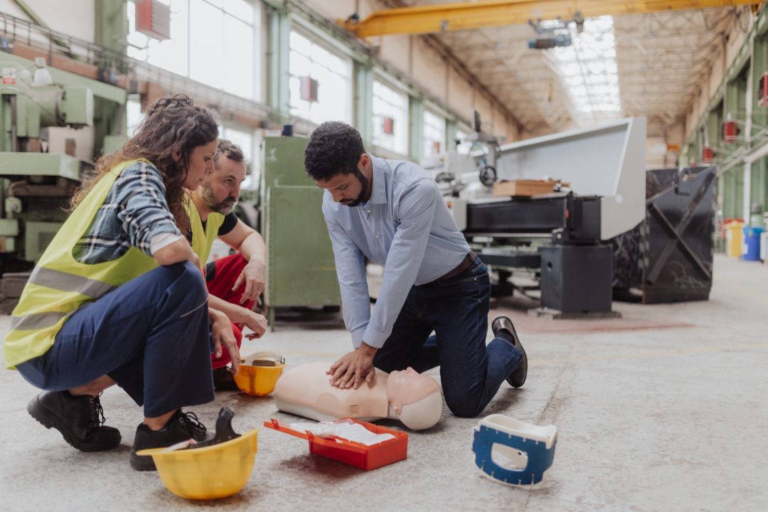 Three people in an industrial workshop perform CPR training on a medical mannequin on the concrete floor.