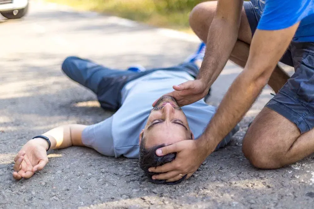 A person kneeling on asphalt stabilizes the head of someone lying unconscious on the ground after an apparent accident.