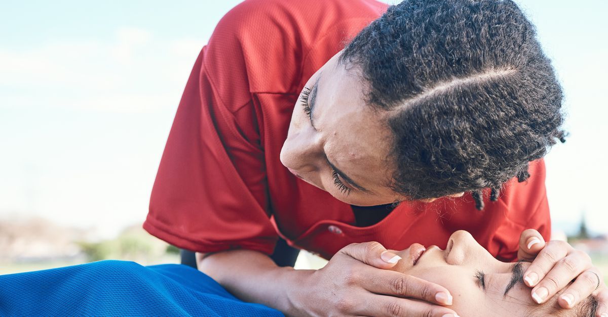 A person in a red shirt checks the breathing and responsiveness of someone lying down outdoors.