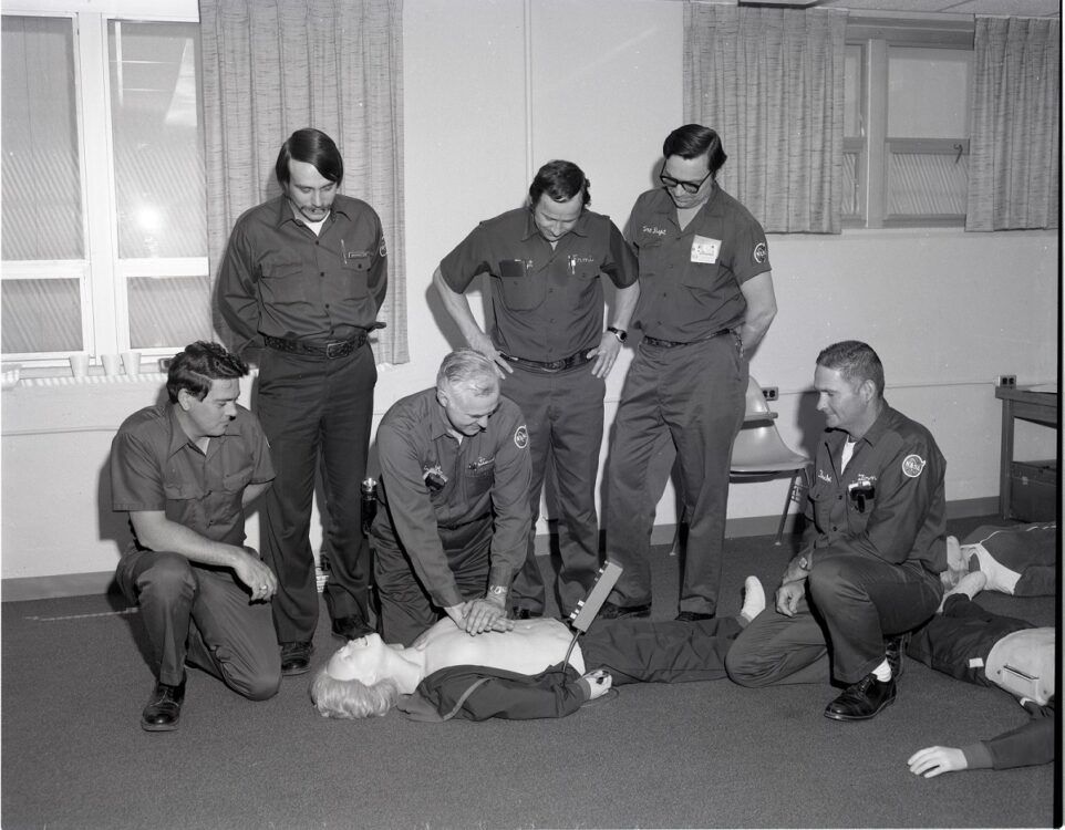 A group of uniformed professionals observe a CPR training session on a medical mannequin in a classroom setting.