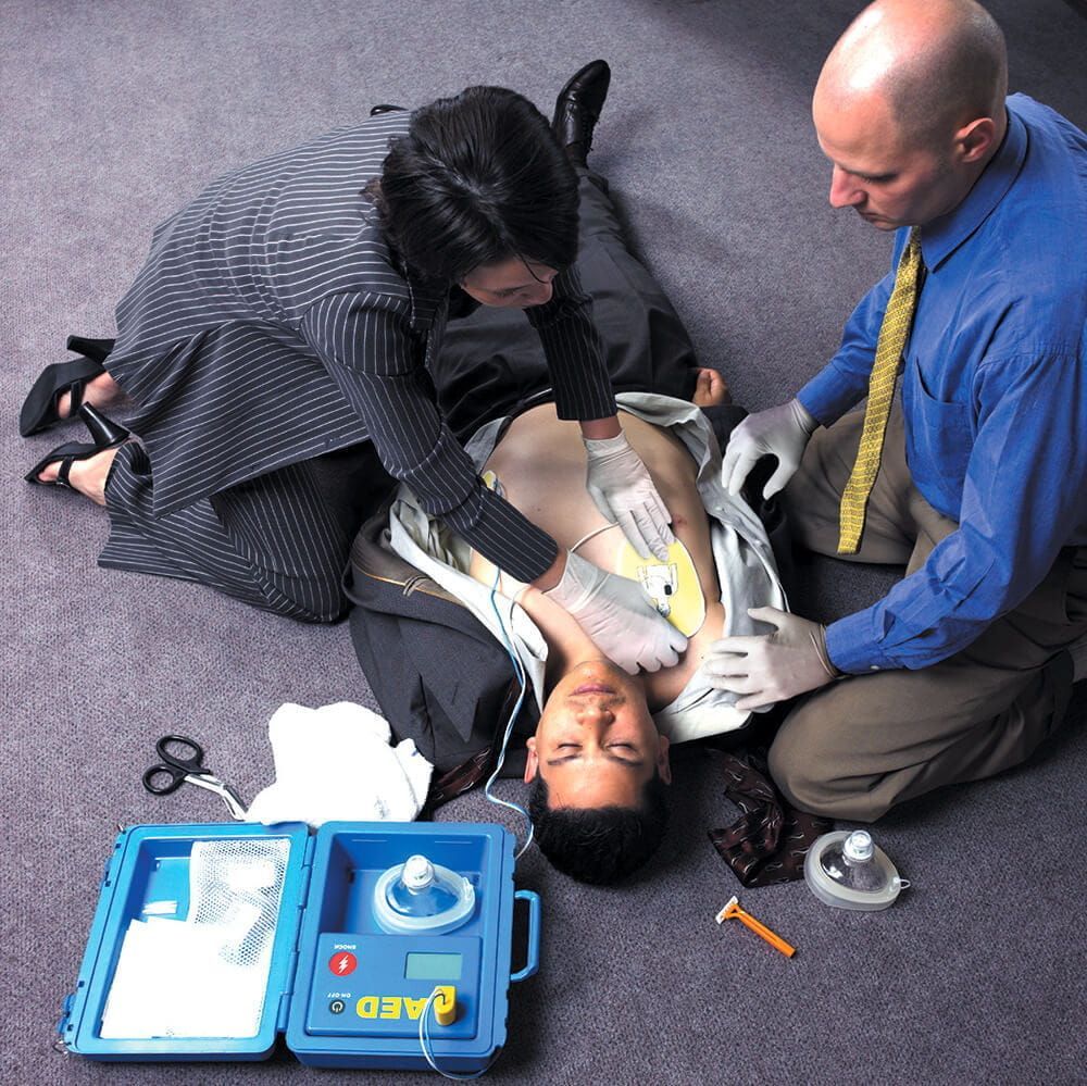Two people in business attire applying AED electrode pads to the chest of a person lying on the floor during a rescue.