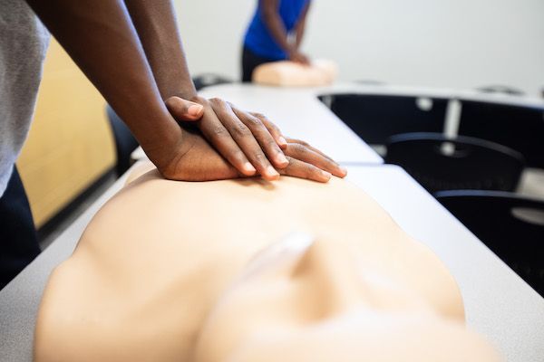 A person performs CPR chest compressions on a medical training mannequin in a classroom setting.