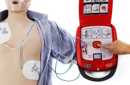 A hand presses the red shock button on a portable AED connected to a training mannequin.