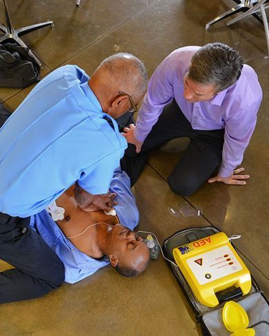 A person in a blue shirt performs CPR on a manikin, with an observer nearby and a yellow AED device on the floor.