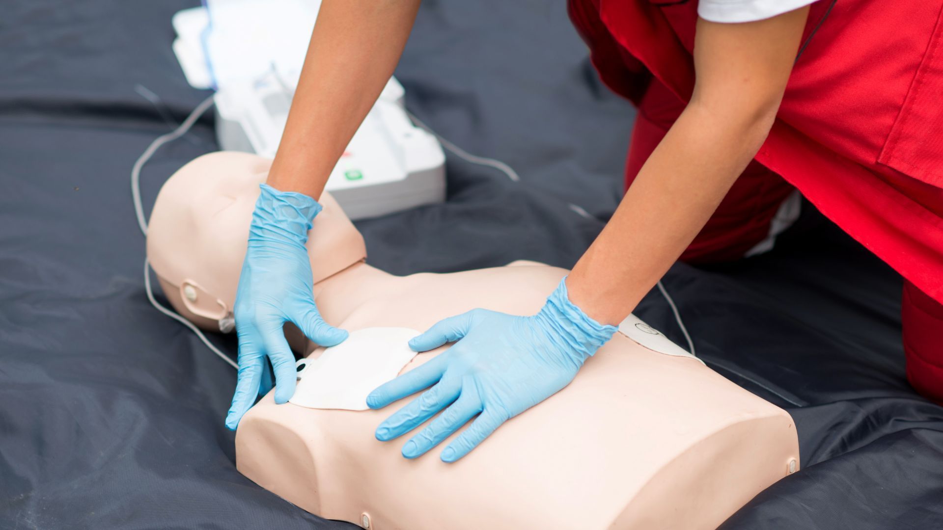 A person in a red uniform and blue gloves places AED pads on the chest of a medical training mannequin.