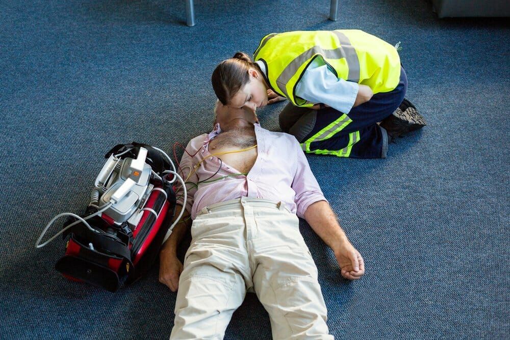 A person in a high-visibility vest performs CPR on a person lying on the floor, with medical equipment nearby.