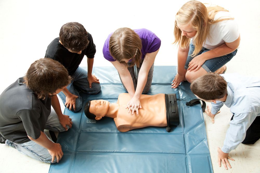 A person performs chest compressions on a CPR mannequin while four others observe on a blue mat in a bright setting.