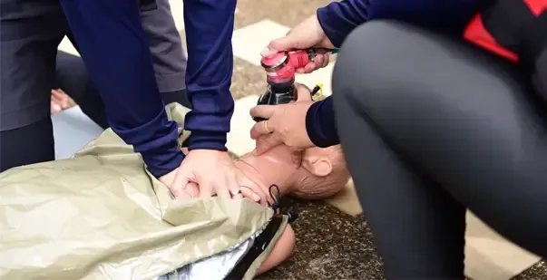 Two people perform CPR training on a mannequin, one administering chest compressions while the other secures a face mask.