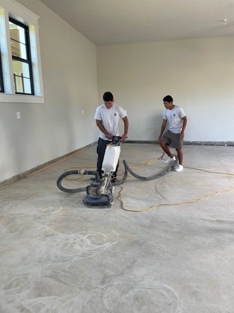 Two workers use a floor grinding machine on a concrete floor in an unfinished room with a window.