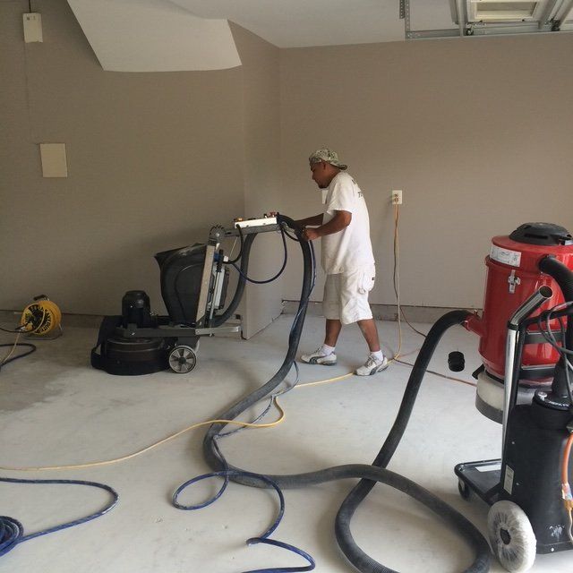 A worker uses a large floor grinder and vacuum system to sand a concrete floor in a neutral-toned room.