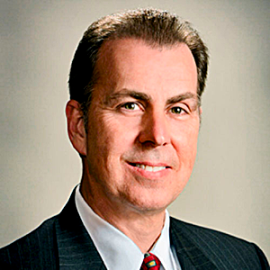 Professional headshot of a smiling man in a dark suit and red tie against a neutral background
