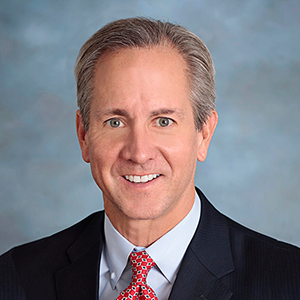 Formal portrait of a man in a dark suit and red patterned tie, smiling against a blue-gray background