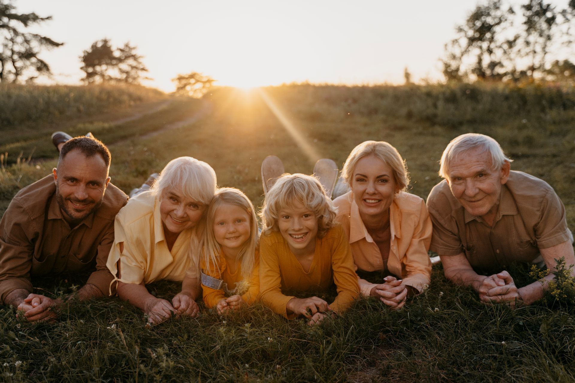 Six people lying in grass at sunset, smiling toward the camera.