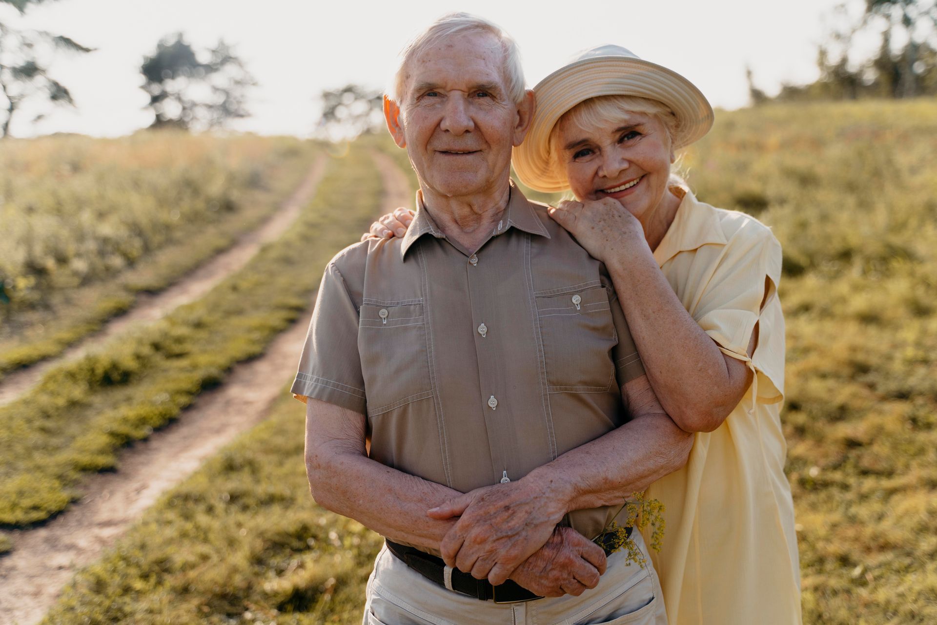 Two people standing together in a sunlit field, smiling and embracing near a dirt path