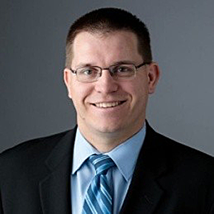 Smiling man in glasses wearing a suit and blue tie against a gray background