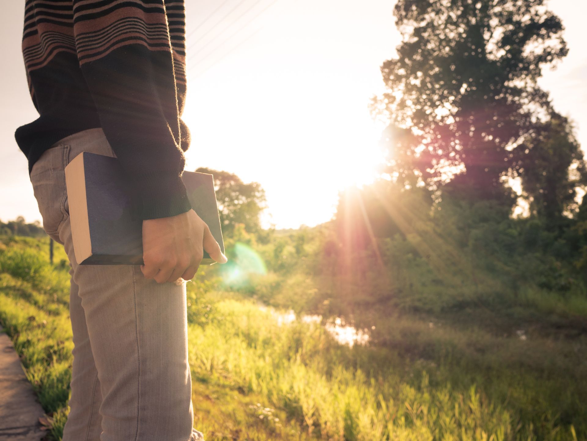 Person holding a book or notebook in a sunlit field at sunset, with trees and water in the background