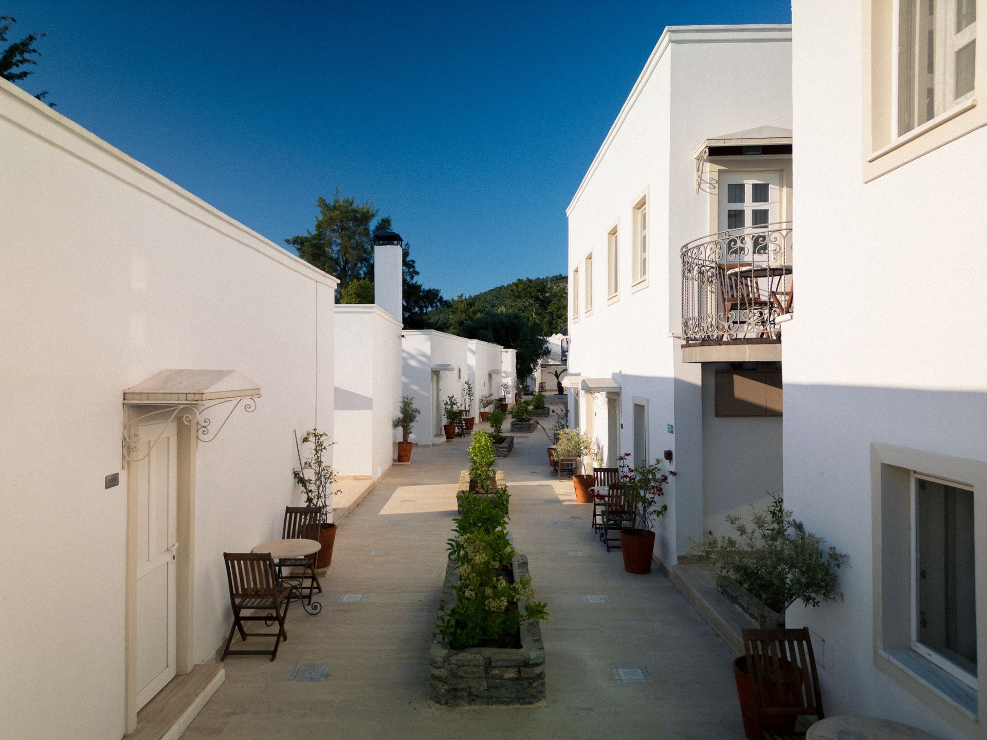 Manzara Boutique Hotel |White courtyard walkway between white buildings, with potted plants and a bright blue sky.
