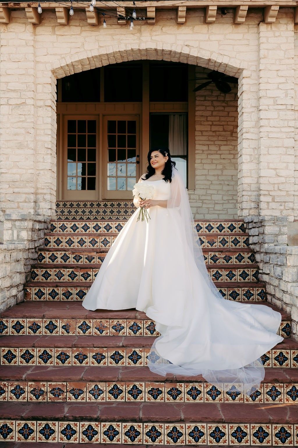 A bride in a white dress is standing in a hallway holding a bouquet of flowers.