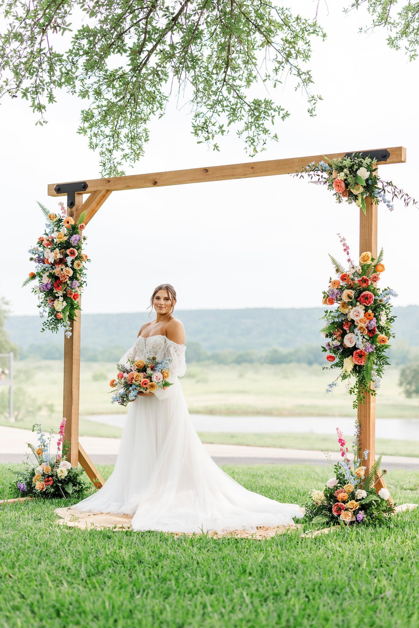A bride in a wedding dress is standing under a wooden arch decorated with flowers.