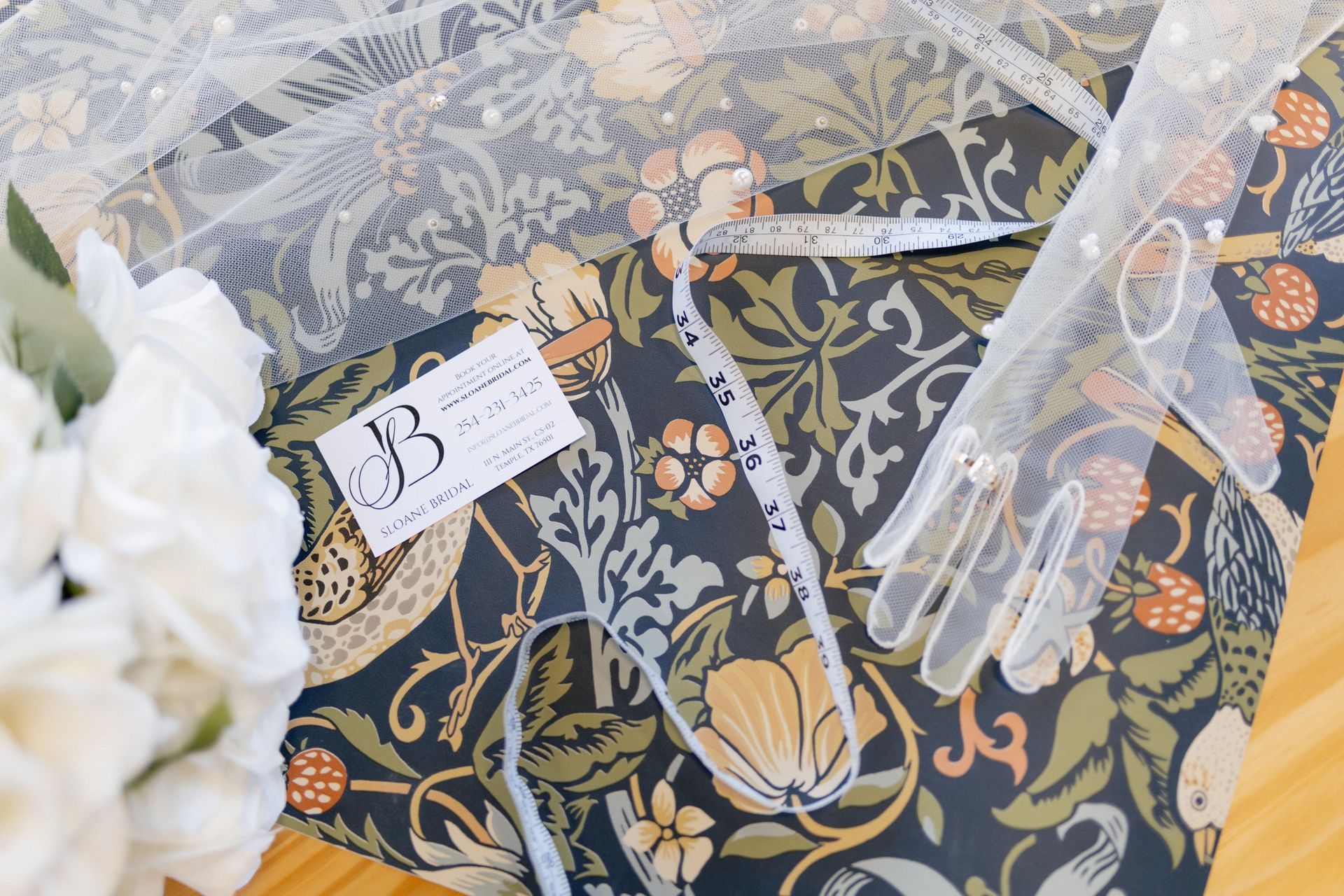 White flowers and lace over a floral-patterned background with a small monogram tag and ribbons