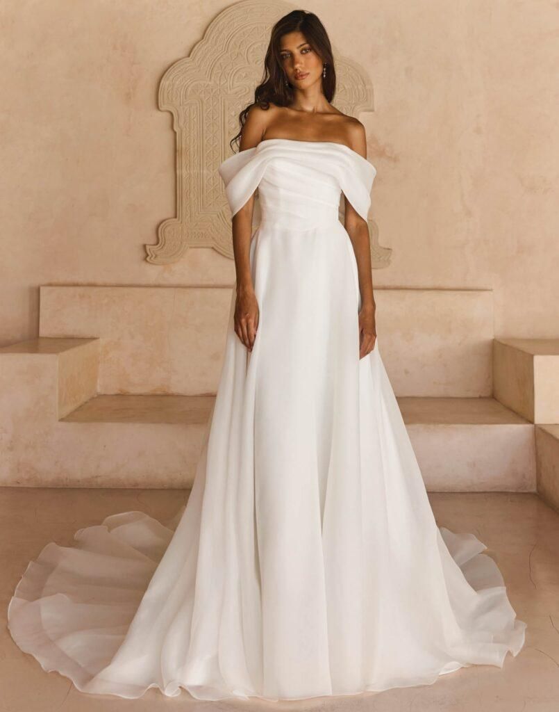 Woman in a white off-the-shoulder gown standing on steps against a beige wall.