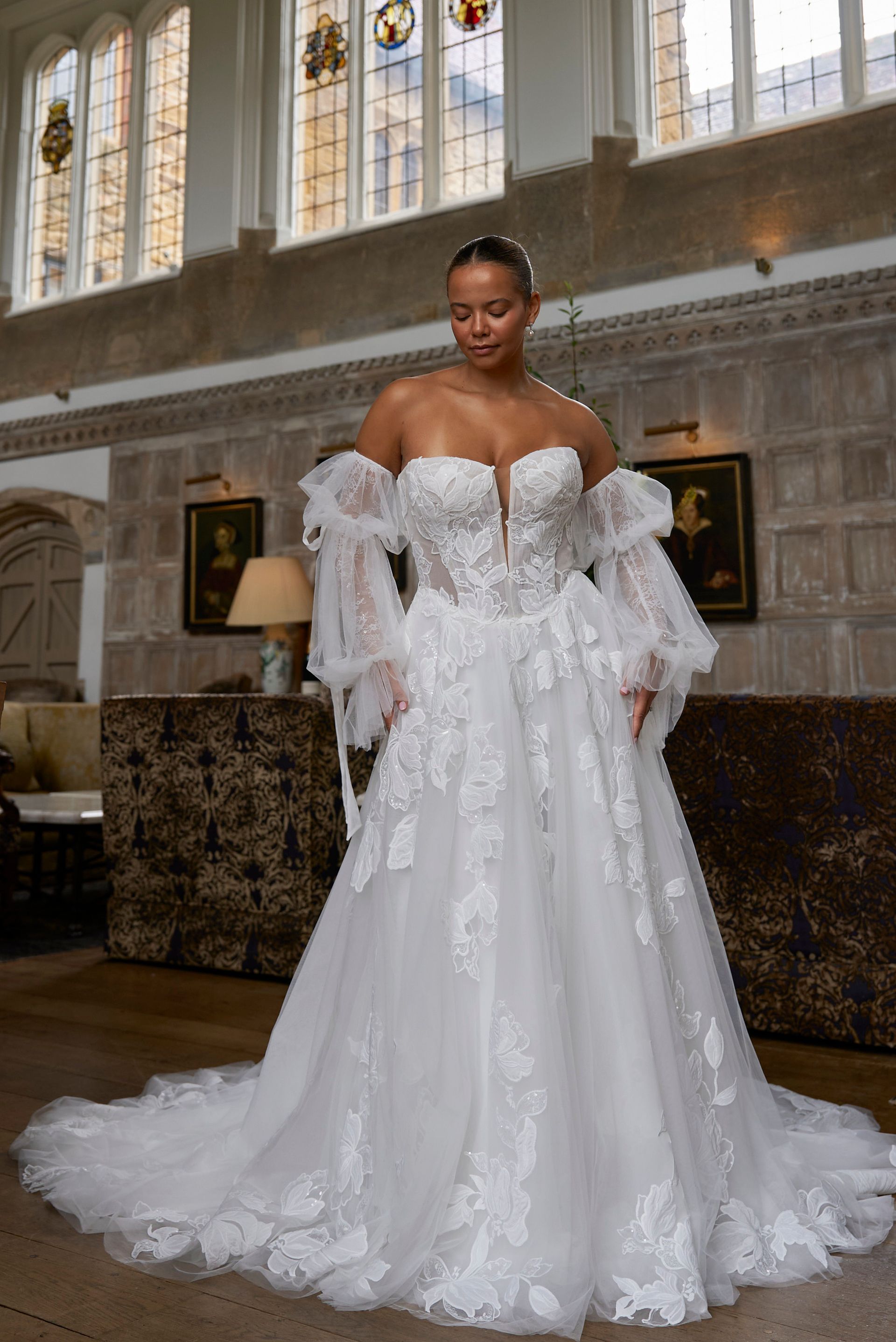 Woman in a white lace ball gown standing in an ornate hall with tall windows.
