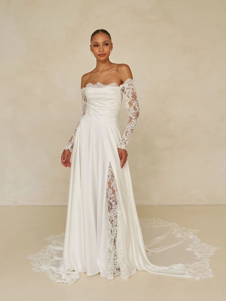 Woman in a white lace bridal gown with a long train, standing against a beige backdrop.