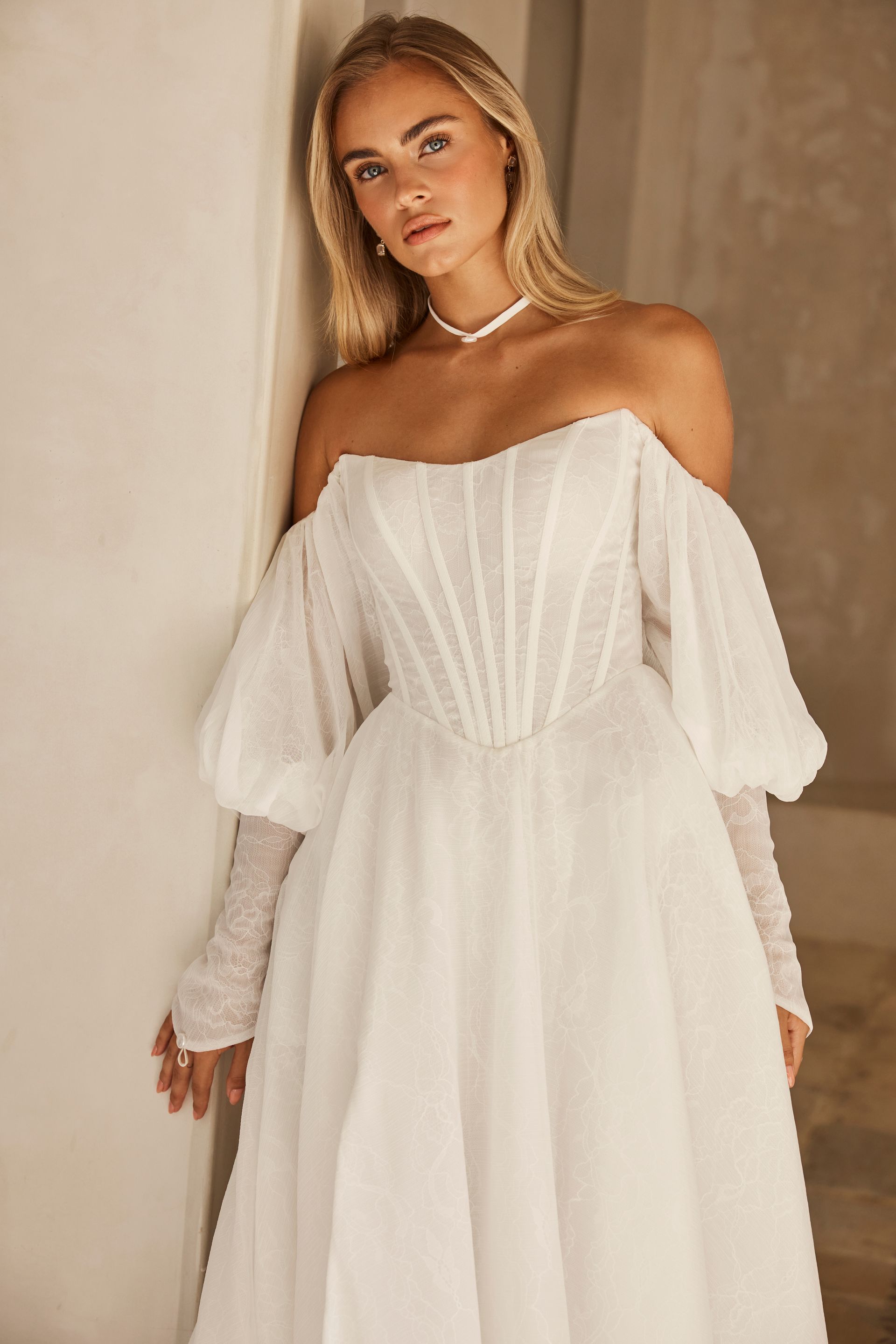 Woman in a white off-the-shoulder dress standing indoors against a beige wall.
