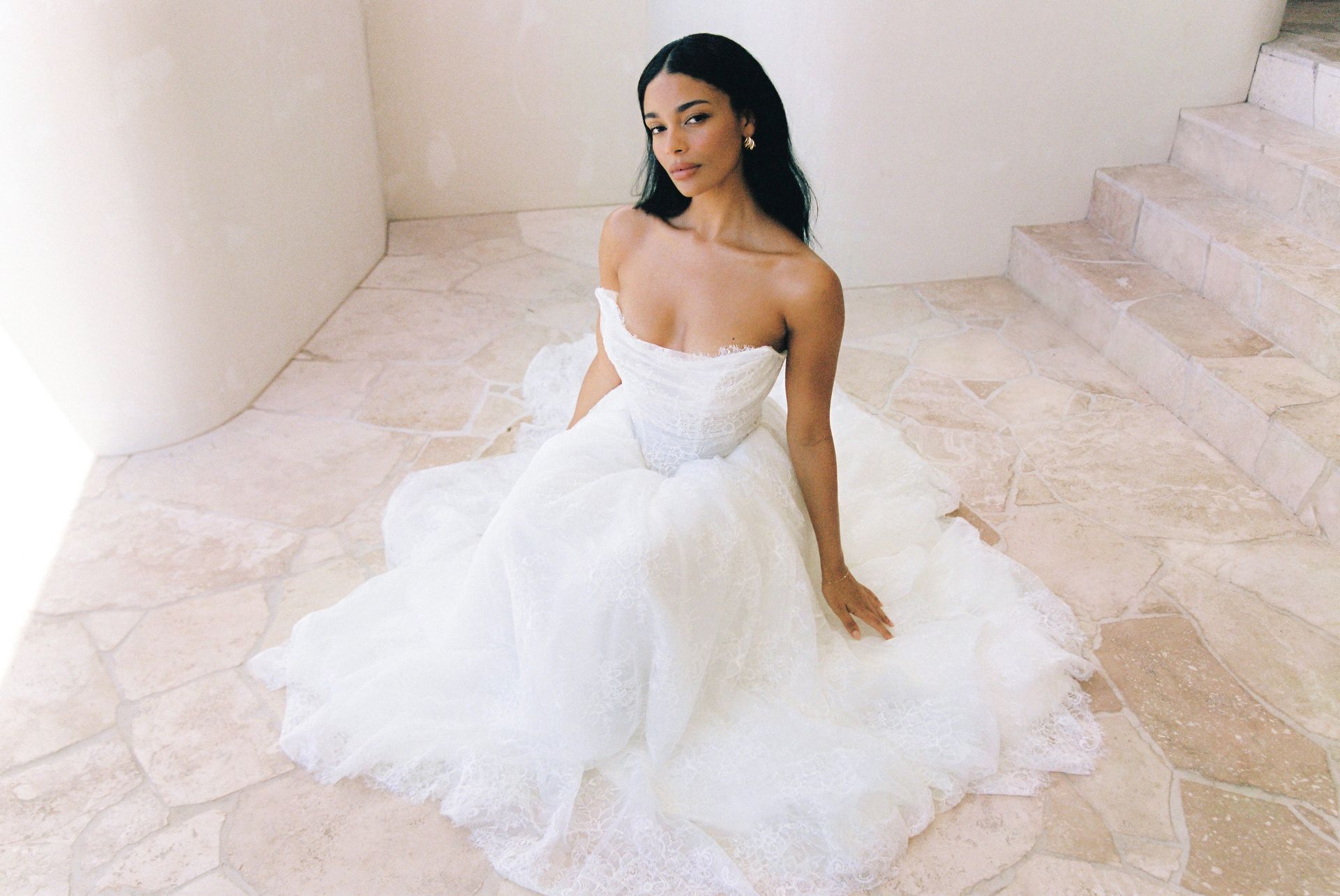 Woman in a white strapless gown seated on a pale stone floor, looking toward the camera.