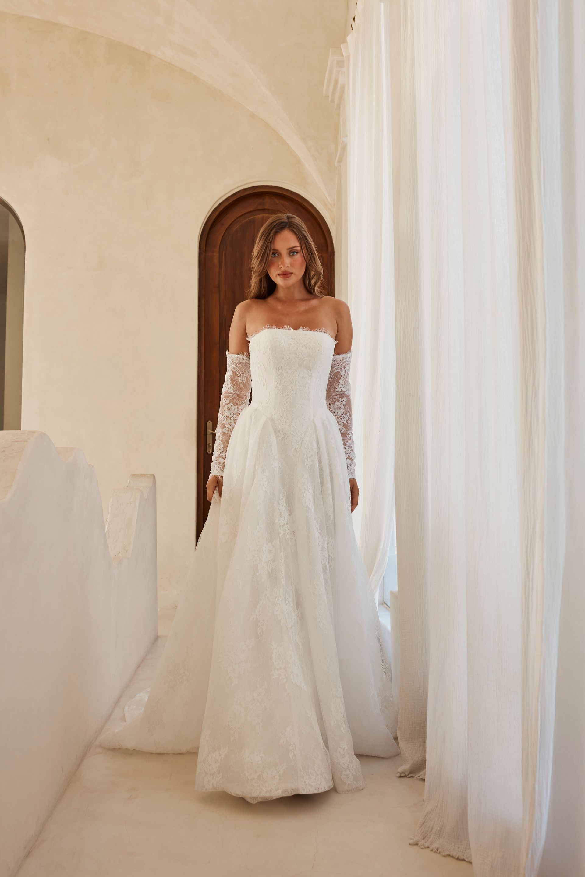 Woman in a white strapless gown standing in a bright arched hallway with flowing sheer curtains