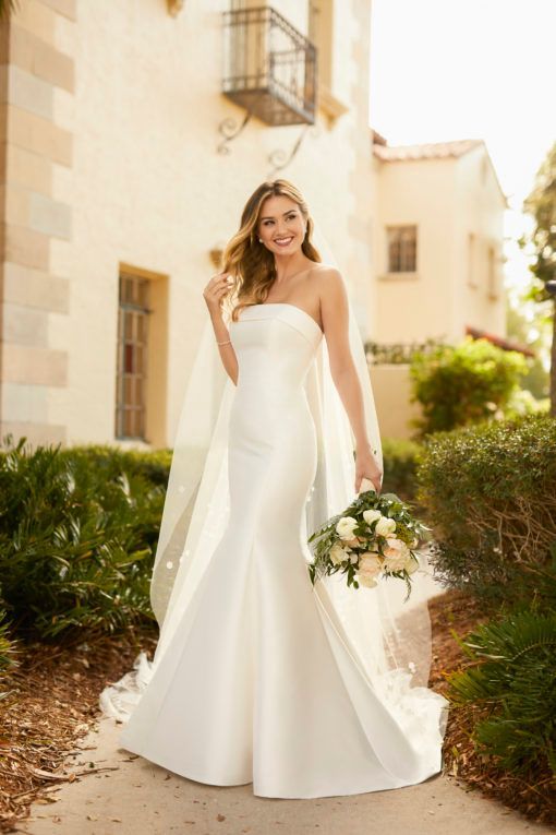 Bride in a white gown holding a bouquet, standing outdoors by a sunlit building