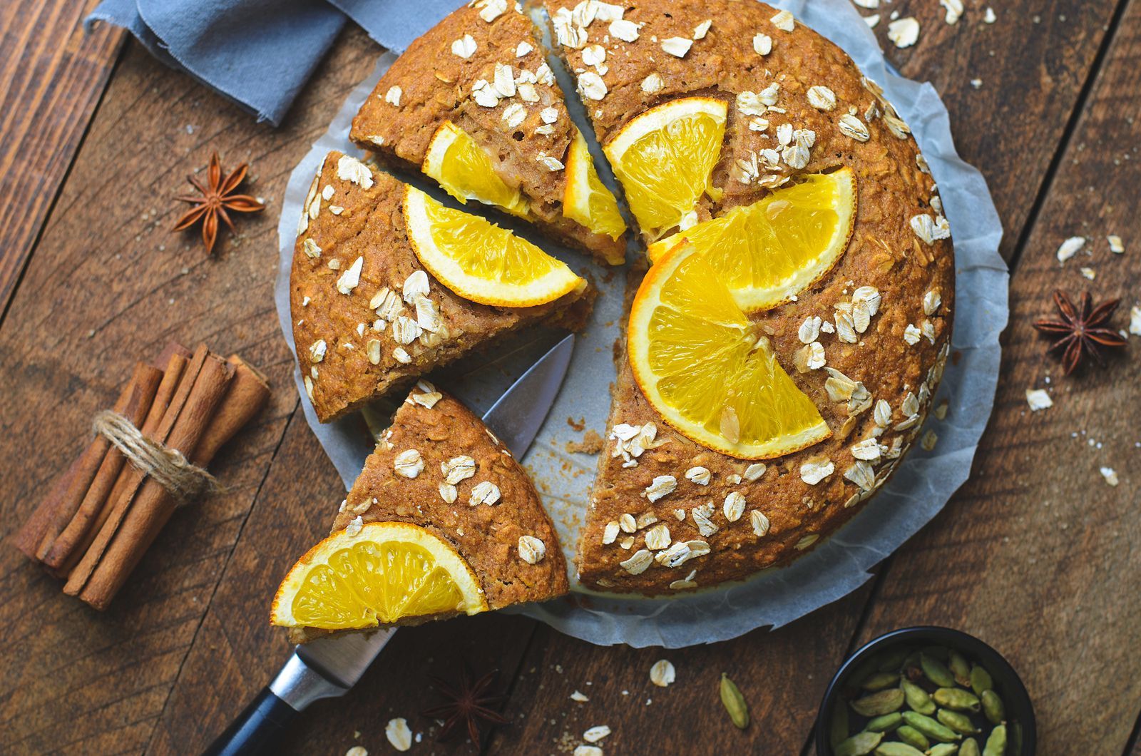 A cake with orange slices and oats is being sliced on a wooden table.