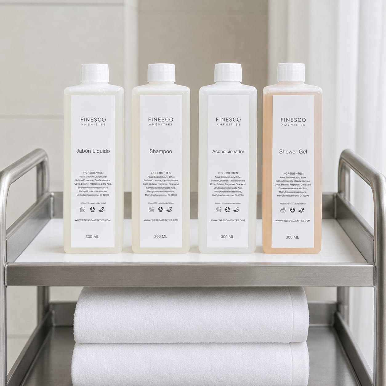 Cuatro botellas blancas sobre una repisa de metal con toallas enrolladas debajo en un baño luminoso o un ambiente de spa.
