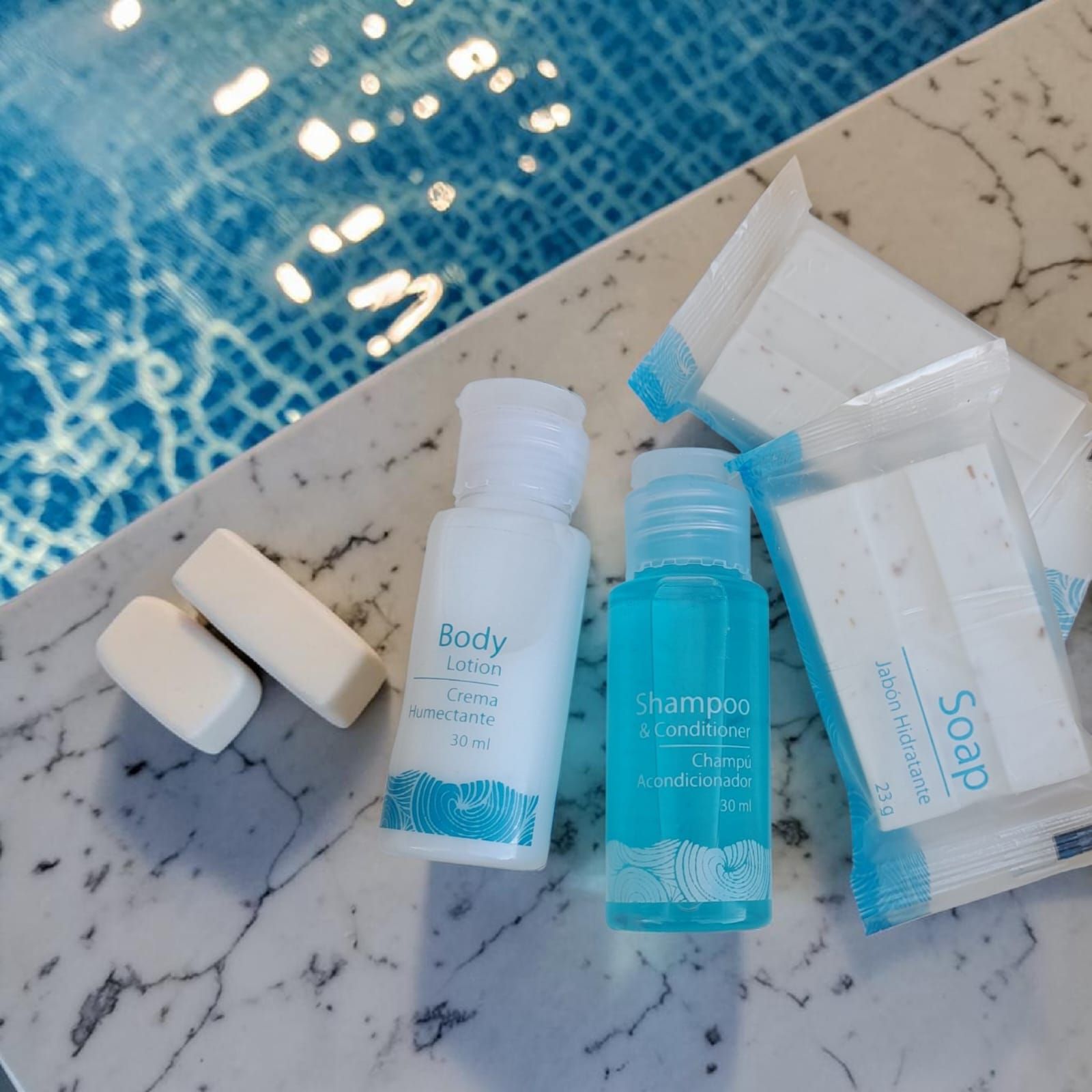 Pequeños frascos de artículos de tocador y pastillas de jabón junto a una piscina azul sobre una superficie de mármol.