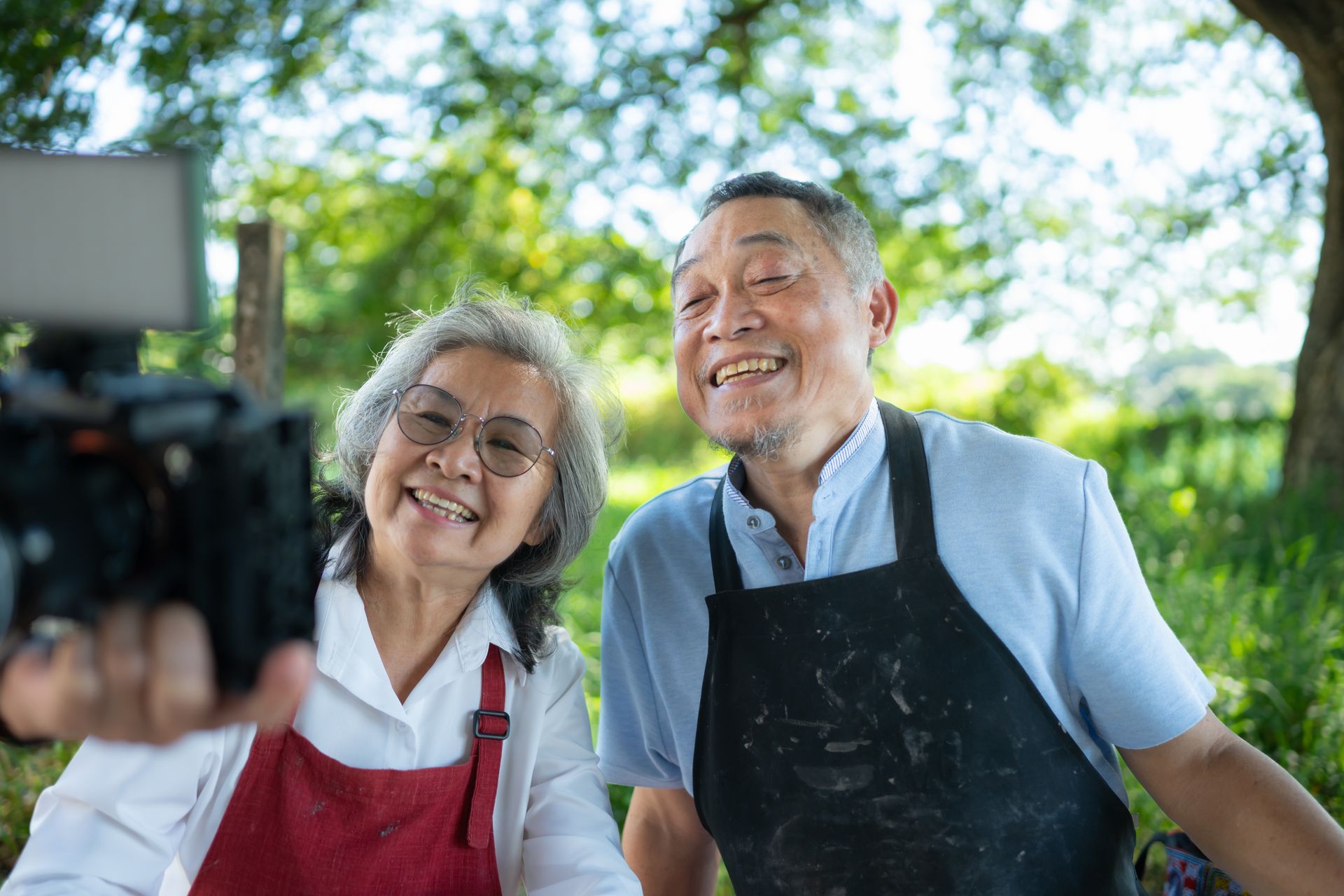 Smiling couple taking a selfie with a camera outdoors, wearing aprons. Green background.