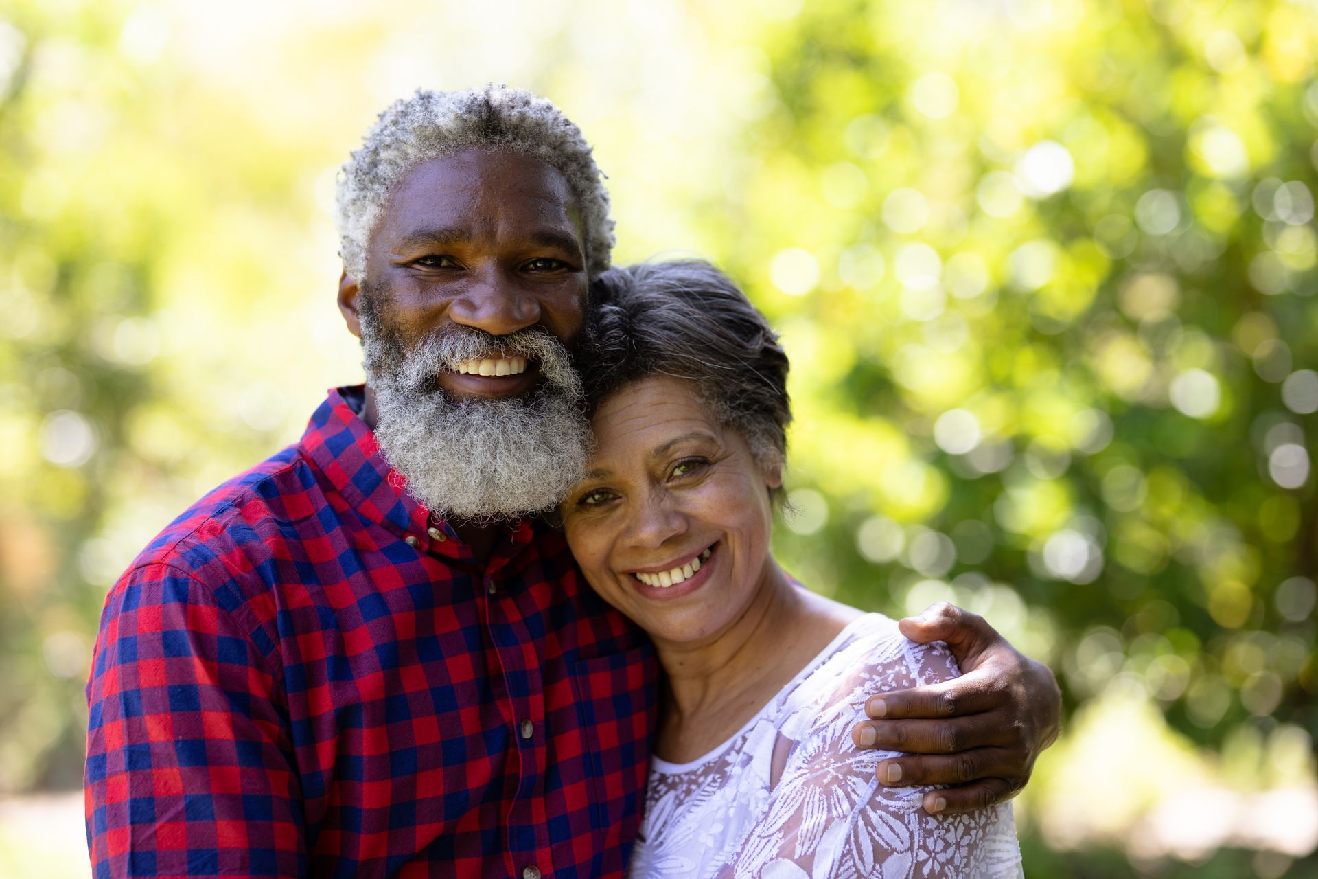 Smiling couple embracing outdoors. Man with gray beard wearing red plaid shirt, woman with lace top. Green background.