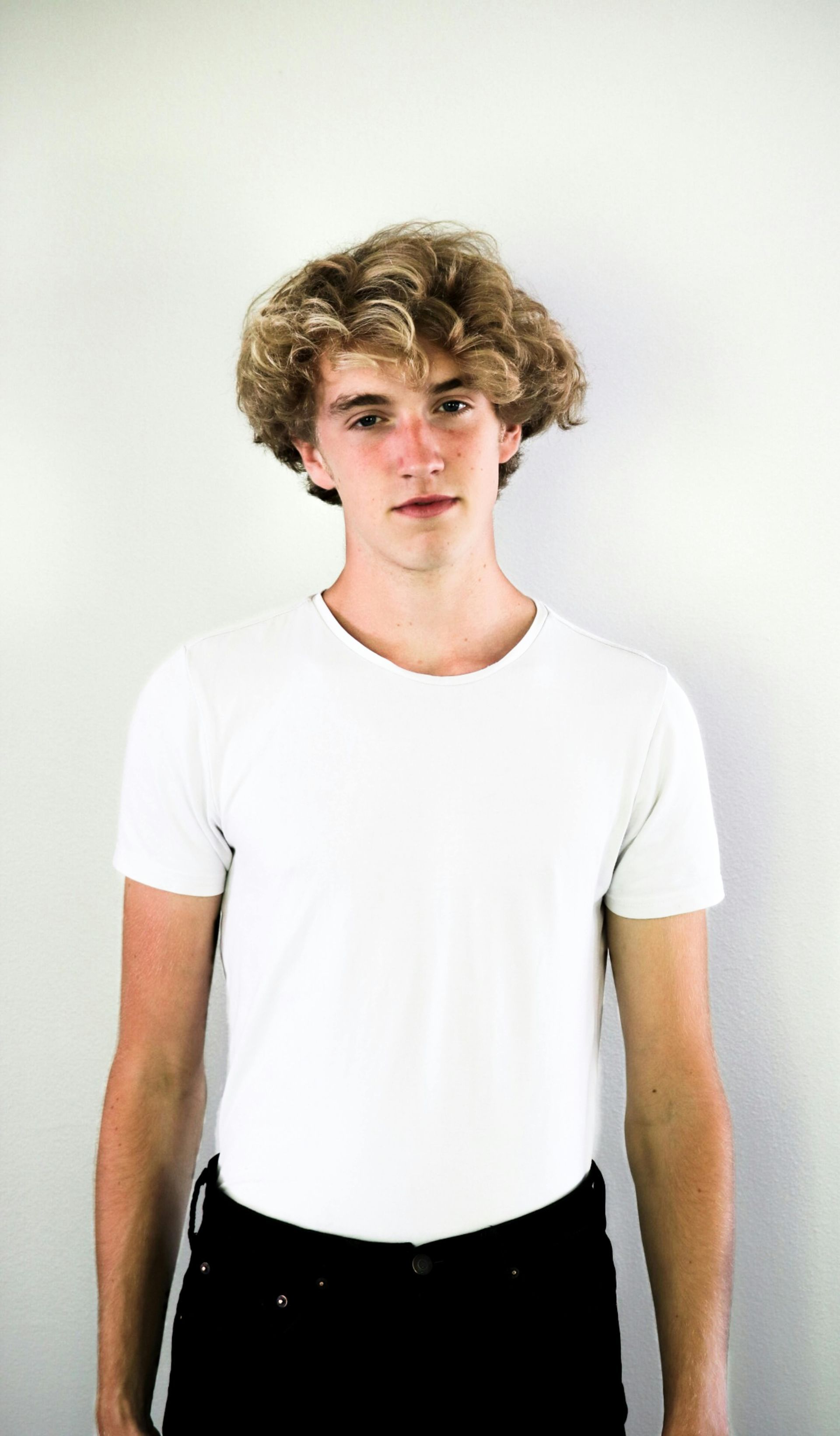 A young man with curly hair is wearing a white t-shirt and black jeans.