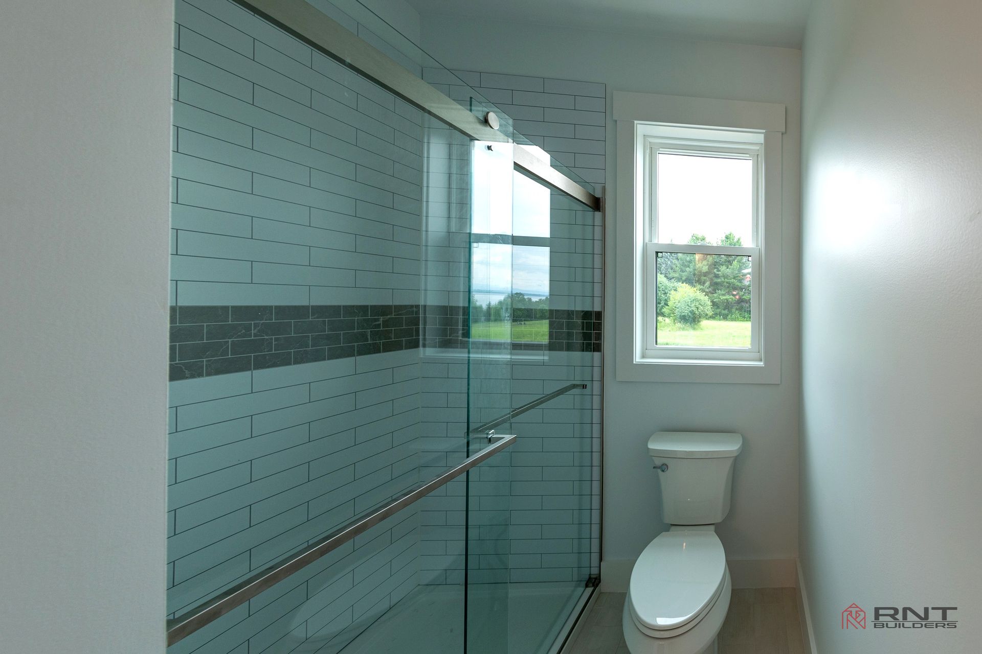 Modern bathroom featuring a glass shower, white tiles, and a toilet near a window overlooking a green landscape.