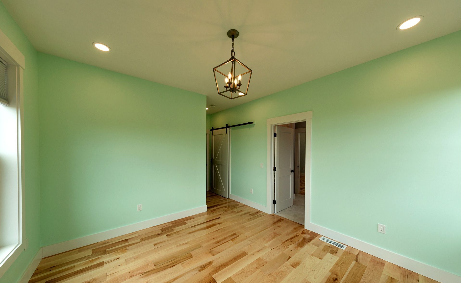 A light-green room with hardwood floors, white trim, and a decorative light fixture; a hallway and doorway are visible.