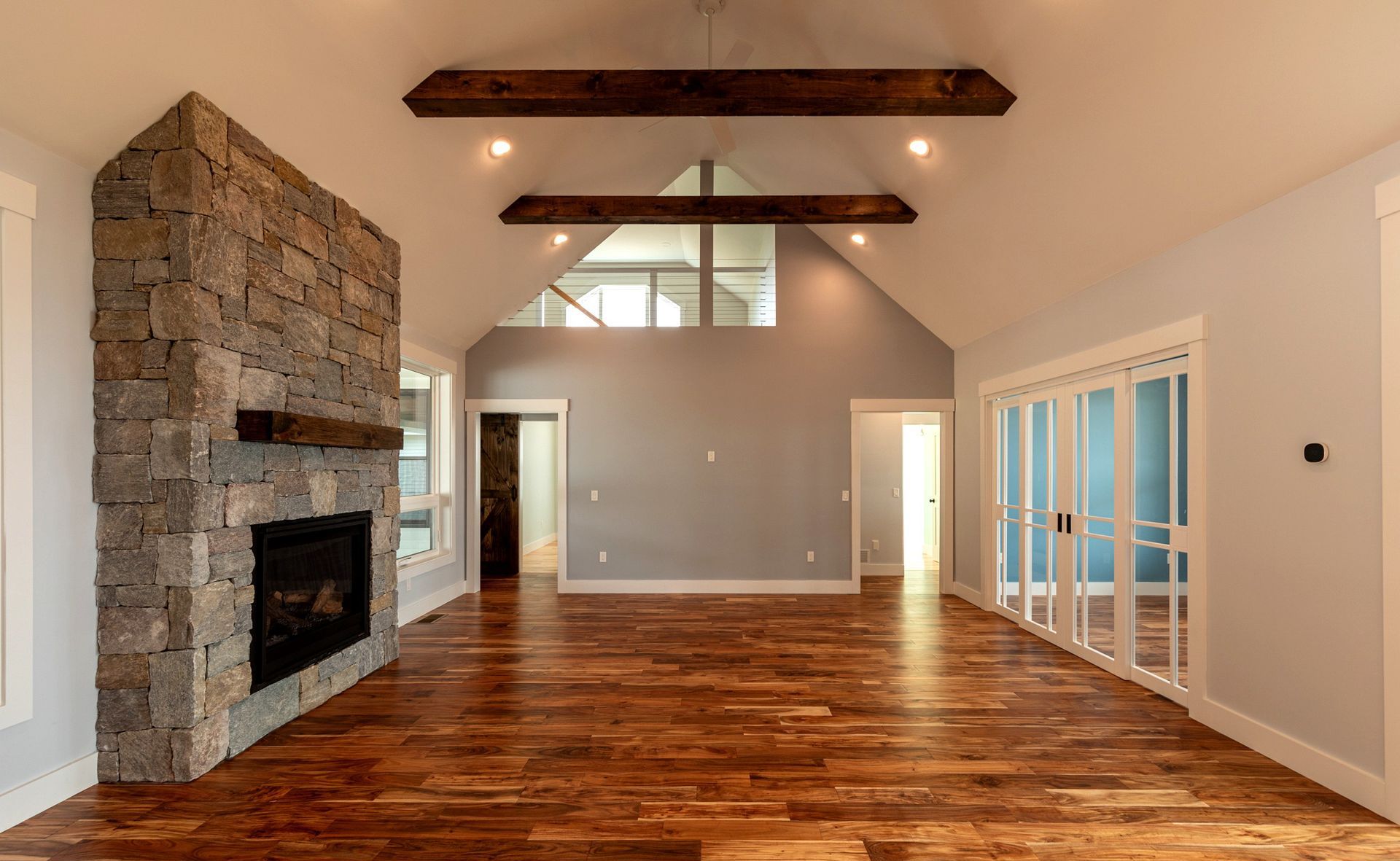 A spacious living room with hardwood floors, a stone fireplace, and exposed wooden beams. Soft blue walls and large windows provide natural light.