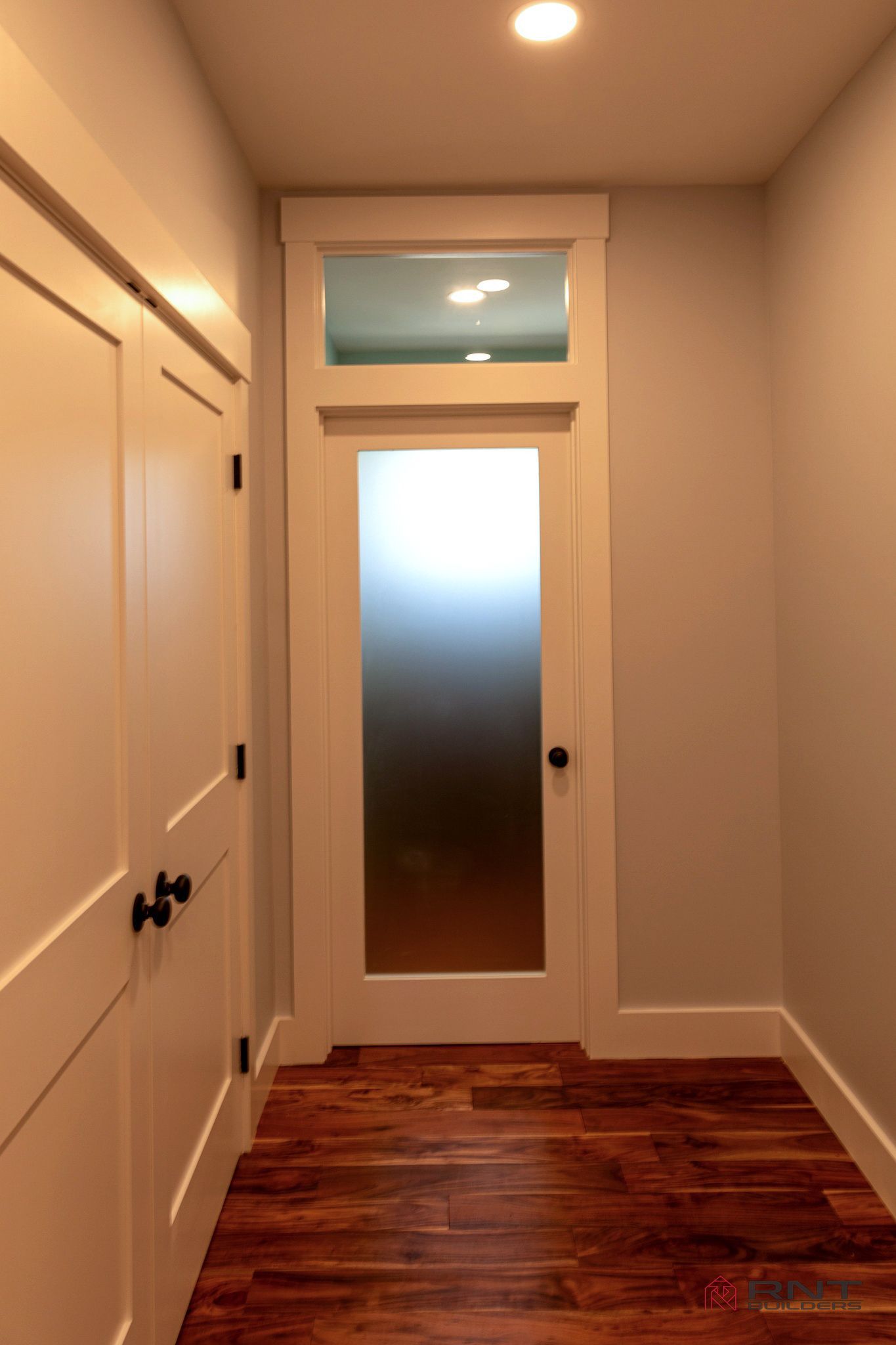 Narrow hallway with a door and frosted glass panel, wood floor, and white trim. A closet door is on the left.