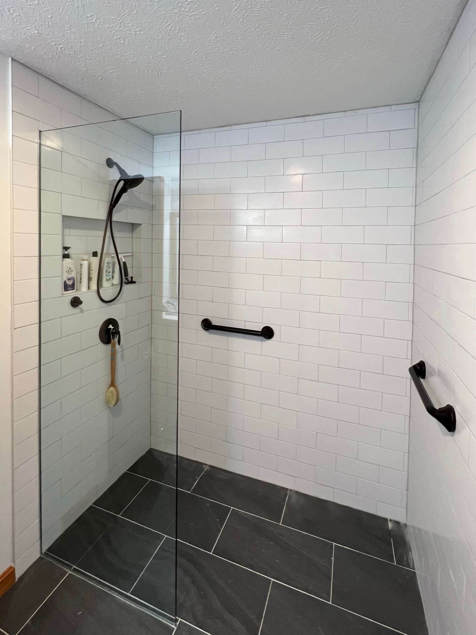 Modern accessible shower with glass door, white tiled walls, black floor, grab bars, and shower niche with toiletries.