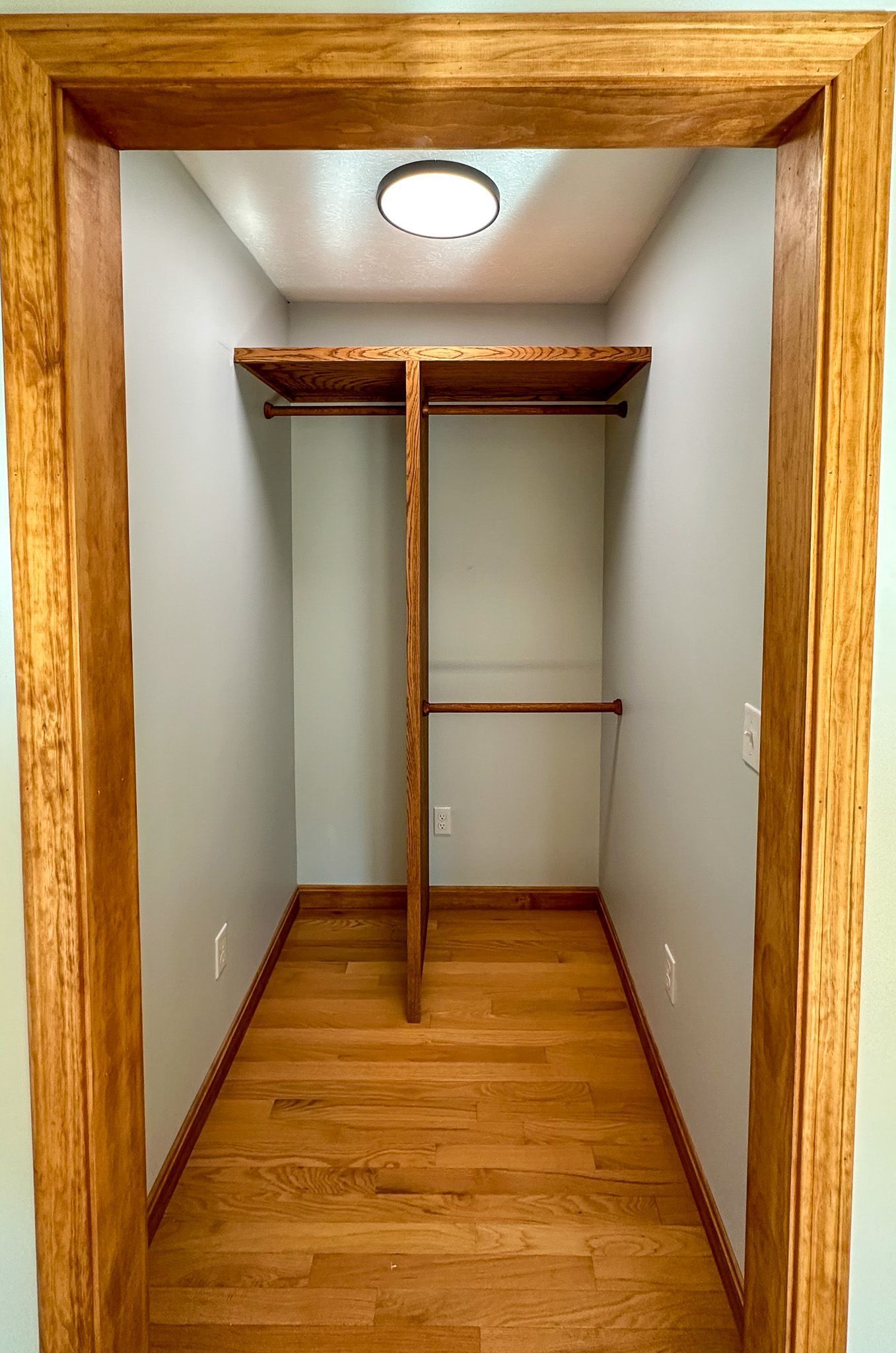 Walk-in closet with light-colored wooden floor and shelving. Pale gray walls and wooden trim. A light fixture is in the ceiling.