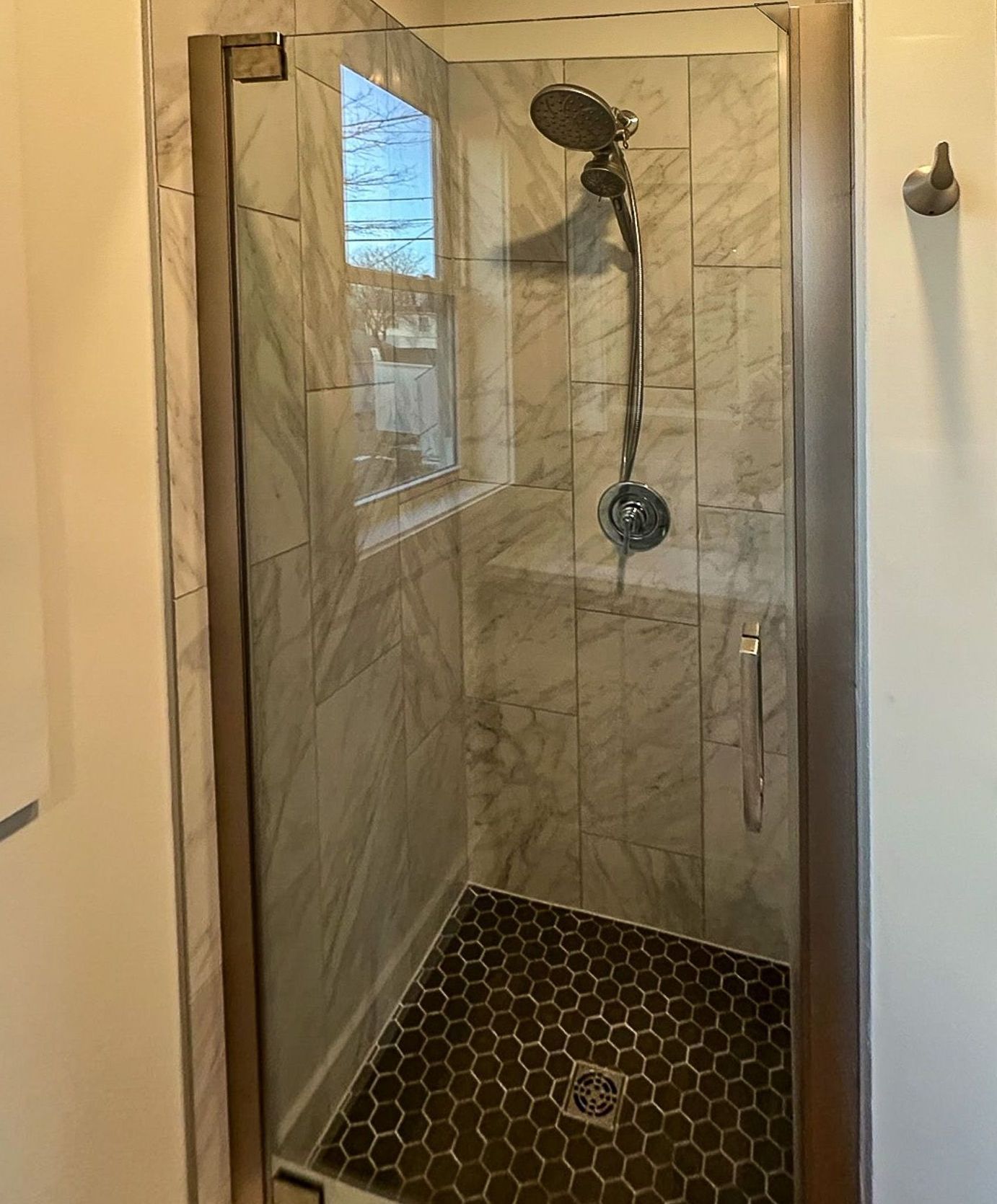 A modern shower with a glass door, marble-like tiles, and a dark hexagonal floor.