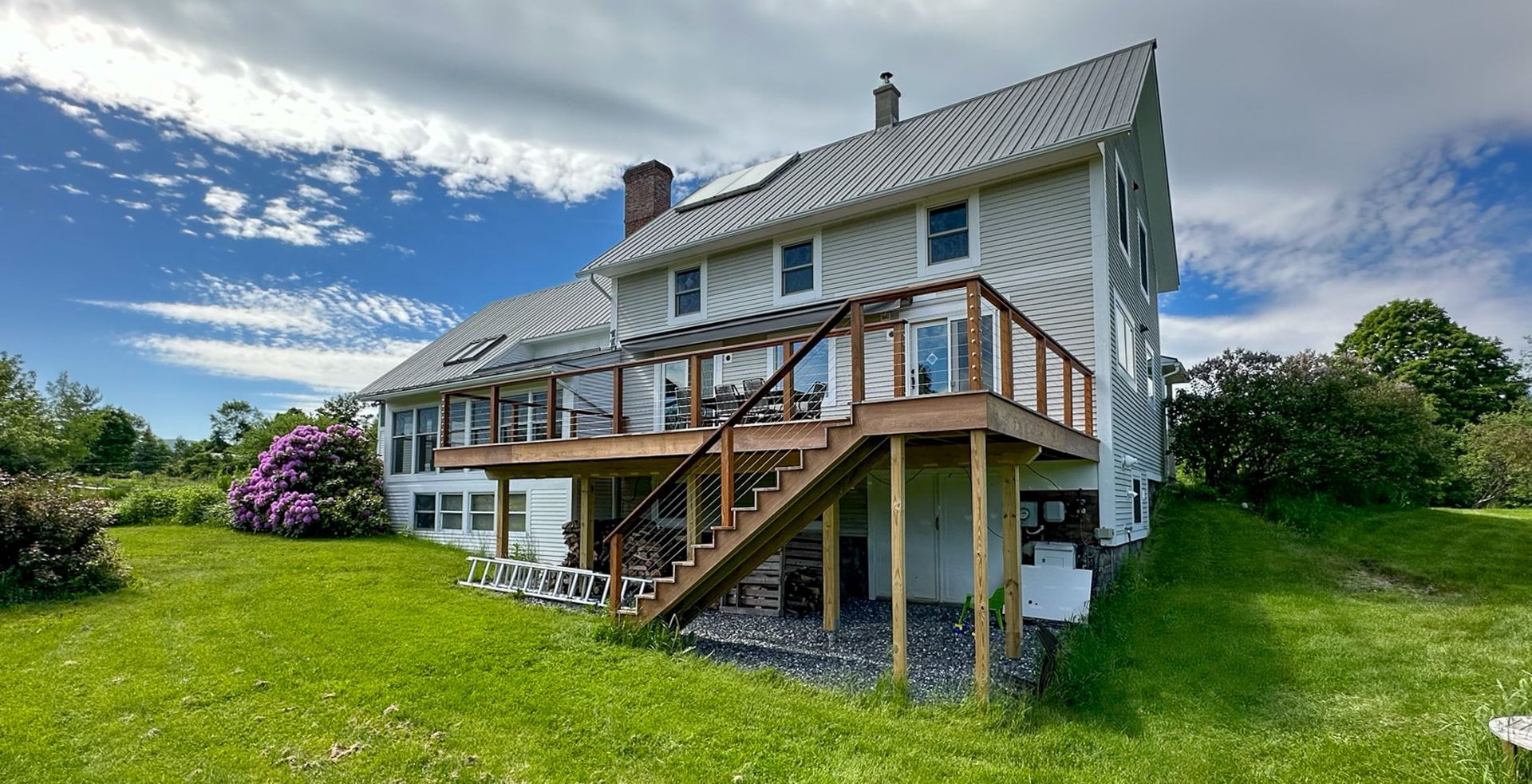 Two-story white house with a large wooden deck and stairs, set on a green lawn under a blue sky with clouds.